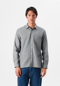 Camisa de botón de manga larga gris con cuello, tela texturizada y un bolsillo en el pecho. Presenta un pequeño logo verde en el lado izquierdo.