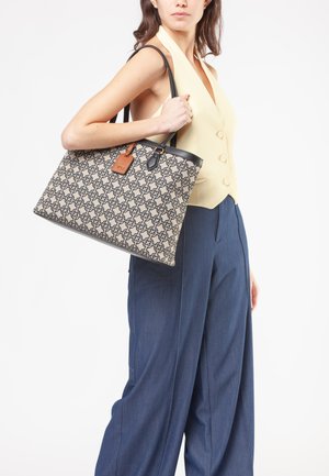 Vrouw draagt een mouwloos beige gilet en blauwe broek en draagt een grote patroon draagtas met zwarte handvatten over de schouder.