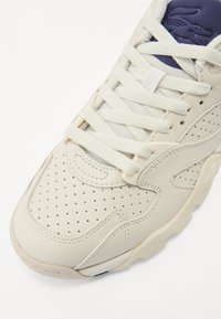Cremfarbene Sneaker mit perforiertem Leder-Obermaterial, strukturierten Akzenten und flachen Schnürsenkeln. Verfügt über eine gepolsterte Sohle und eine marineblaue Zunge.