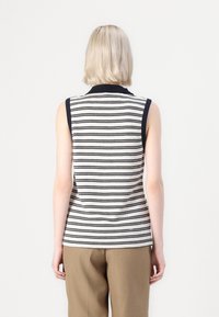 Mouwloos poloshirt in crème met marineblauwe strepen, met een gebreide textuur en een effen gekleurde kraag. G gedragen met een paar bruine broeken.