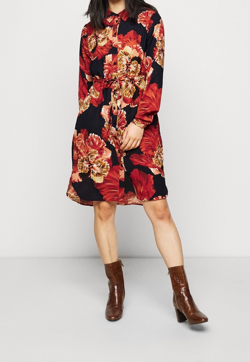 Abito floreale in tessuto scuro, con grandi fiori rossi e dorati, maniche lunghe, chiusura con bottoni e una cintura in vita. Stivaletti marroni con tacco.