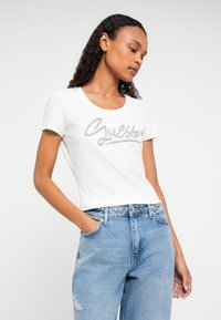 Frau, die ein weißes Guess-Logo-T-Shirt und hellblaue, zerrissene Jeans trägt, steht mit einer Hand in der Tasche vor einem einfarbigen Hintergrund.