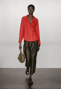 Rød linned skjorte med knapper, brune brede bukser, sorte sandaler og en lille tan taske. Model går på en ensfarvet baggrund.