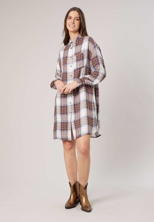 Femme souriante, portant une robe chemise ample à manches longues à carreaux avec des boutons et des bottines marron métalliques, debout devant un fond uni.