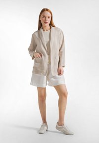 Blazer in lino beige chiaro con colletto a scialle, abbinato a una canotta beige e pantaloncini bianchi. Tessuto liscio, design minimalista e stile casual.