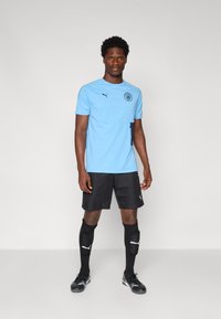 Lyseblå træningsskjorte med korte ærmer, sort logo, parret med sorte shorts og sorte sokker, der har Puma-branding.