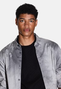 Camicia grigia con colletto e sottili accenti di logo. Indossata sopra una t-shirt nera con texture. Finitura in tessuto liscia.
