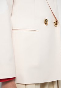 Lichtroze blazer met dubbele goudkleurige knopen, gestructureerde schouders en een subtiel zakdetail. Soepele stof met een getailleerde pasvorm.