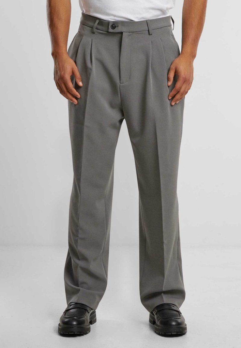 Pantalones gris de plisado, hechos de una tela suave, con cierre de botón, bolsillos laterales y un diseño de pierna recta. Combinados con zapatos negros.