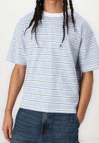 Camisa de manga corta a rayas con rayas en azul claro y blanco, y acentos en negro. Está hecha de algodón suave, con un cuello redondo y un corte relajado.
