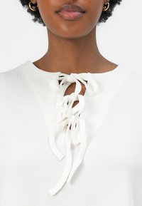 Weißes, strukturiertes Oberteil mit rundem Ausschnitt und Schlüssel-Ausschnitt, versehen mit Bändern. Glattes Gewebe mit dezentem Kräuseldesign.