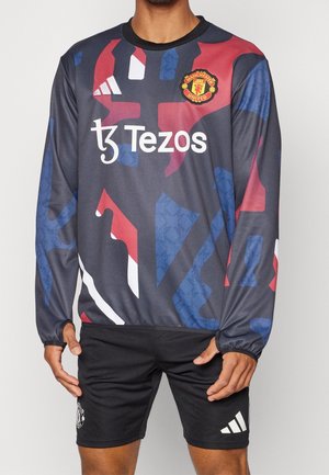 Haut d'entraînement à manches longues de Manchester United avec un motif géométrique noir, rouge et bleu, et le logo du sponsor Tezos sur la poitrine.