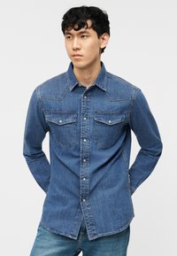 Νέος άνδρας που φοράει μπλε τζιν πουκάμισο με κουμπιά και θήκες στο στήθος, στέκεται με τα χέρια πίσω από την πλάτη μπροστά σε απλό φόντο.