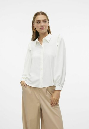 Camisa blanca de botones con cuello, detalles de volantes en los hombros y mangas largas y sueltas. Combinada con pantalones de beige con pliegues.