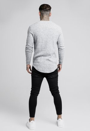 Mężczyzna z tatuażami na szyi i rękach, ubrany w szarą koszulę z długim rękawem, czarne spodnie i białe trampki, stoi odwrócony plecami na jednolitym tle.