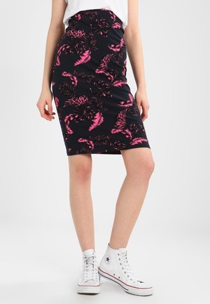 Vrouw met een zwarte rok tot op de knie met roze bladpatronen, wit topje en witte hoge sneakers, staand tegen een effen achtergrond.