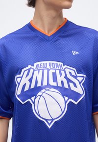Άτομο που φοράει μπλε μπλουζάκι μπάσκετ των New York Knicks με πορτοκαλί περιθώριο και μεγάλο λευκό λογότυπο μπάσκετ στο στήθος.