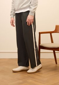 Pantaloni gris leggero con due strisce bianche laterali, vestibilità rilassata, realizzati in tessuto morbido. Abbinati a sneaker beige su un pavimento di legno.