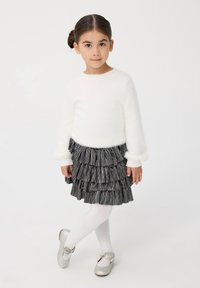 Maglione bianco soffice con maniche lunghe, gonna a strisce nere e grigie con volant, collant bianchi e ballerine argentate lucide con dettagli a fiocco.