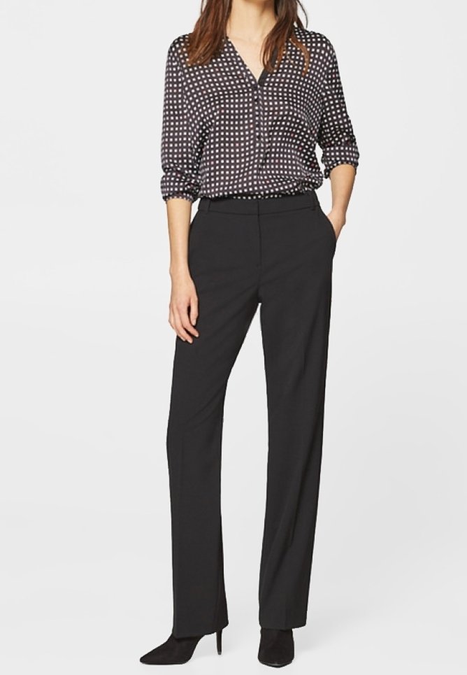 Kvinna klädd i svarta skräddarsydda byxor, svarta spetsiga klackar och en svart blus med vitt rutmönster, stående med en hand i fickan.