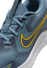 Nike Cosmic Runner in blue with yellow accents. Features mesh upper, rubber sole, and flat laces. Modern design with textured materials.