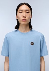 Světlounce modré bavlněné tričko s krátkým rukávem a kulatým výstřihem. Obsahuje kulatou nášivku s logem na levé části hrudi. Měkká textura.