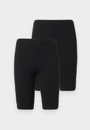 Kobieta z kręconymi blond włosami, ubrana w czarną bluzkę z długim rękawem, czarne spodenki do kolan, białe skarpetki i czarne sznurowane buty, stojąca na białym tle.