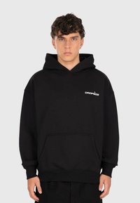 Giovane uomo con capelli ricci, che indossa un hoodie nero con tasca frontale e il logo bianco "DROPSIZE" sul petto, in piedi di fronte a uno sfondo semplice.