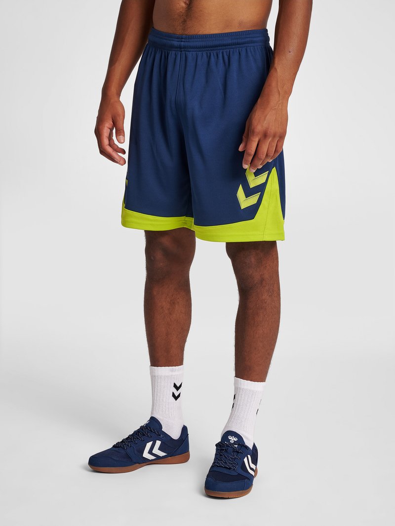 Hombre con pantalones cortos deportivos azul marino y verde neón, calcetines blancos hasta el tobillo y zapatillas deportivas azul marino sobre fondo gris.