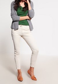 Cardigan grigio, t-shirt verde aderente, pantaloni bianchi slim-fit e scarpe slip-on beige con dettagli decorativi.