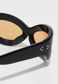 Occhiali da sole neri con lenti arancioni, montatura in plastica spessa, superficie liscia e punte di metallo argentato come dettagli decorativi sulle astine.