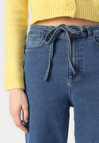 Suéter amarelo tricotado, cropped, com punhos canelados, combinado com jeans azuis de cintura alta, apresentando um cinto de cordão amarrado e detalhes de costura sutis.