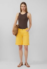 Shorts en denim jaunes avec des poignets retroussés, associés à un débardeur marron ajusté. Tenant un sac en tricot. Sandales avec une sangle à motif léopard.