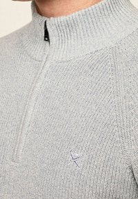 Primo piano di un maglione a maglia grigio chiaro con zip a metà e un piccolo logo ricamato di mazze da golf incrociate sul petto.