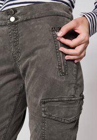 Graue Cargo-Hose mit glatter Textur, ausgestattet mit einer Reißverschlusstasche und zwei Nieten am Bund, die ein lässiges Design und funktionale Details präsentieren.
