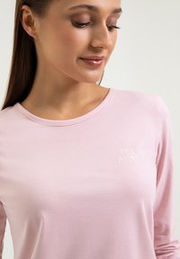 Różowa koszula z długim rękawem, wykonana z miękkiego materiału, z okrągłym dekoltem i białym haftowanym logo na piersi. Gładka tekstura.
