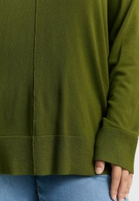 Olivgrüner Strickpullover mit gerippten Bündchen und Saum, der eine Mittelnaht und eine weiche, glatte Textur aufweist. Kombiniert mit hellblauen Jeans.