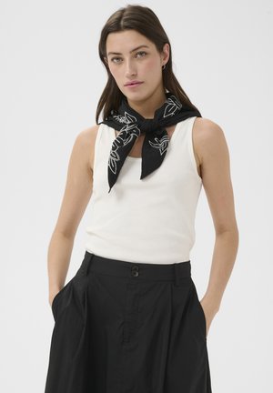 Naine, kellel on seljas valge varrukateta särk, mustad kõrge vöökohaga püksid ja must mustriga sall kaelas, käed taskutes.