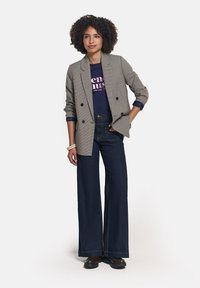 Blazers gris con patrón sobre una camiseta gráfica azul marino, combinados con jeans oscuros de campana. El aspecto cuenta con solapas anchas y detalles de costura en contraste.