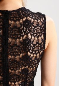 Svart spetsöverdel med blommönster, genomskinliga sektioner och hög halsringning. Synlig dragkedja bak, slät tyg nedtill med en åtsittande silhuett.