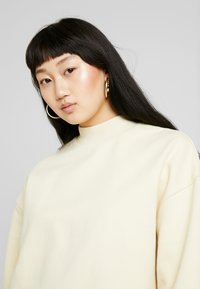 Világos krémszínű pulóver magas nyakkal, laza fazonnal és puha textúrával. Tágas sziluett jellemzi, amelyet nagy arany fülbevalók egészítenek ki.