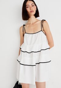 Robe blanche avec des couches superposées et des bordures noires, fines bretelles et une silhouette ample et fluide. Tissu lisse avec une texture douce.