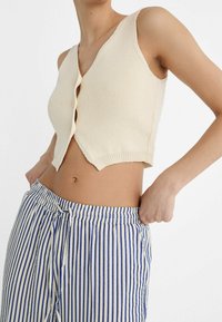 Vrouw in een crèmekleurige gebreide cropped vest en blauw-witte verticaal gestreepte broek, die met één hand de tailleband aanpast.