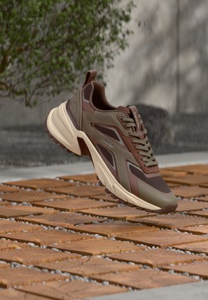 Bruine en beige Calvin Klein sneaker met dikke zool, geplaatst bovenop houten tegels en grind, met groen en een betonnen muur op de achtergrond.