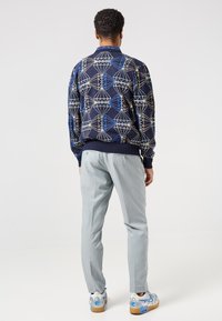 Man wearing blue geometric patterned sweater, light grey trousers, and white sneakers with blue accents, standing with back to camera.