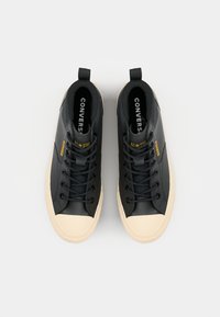 Baskets montantes noires en caoutchouc et textile, avec des lacets noirs, un bouts en crème et un logo jaune sur le côté.