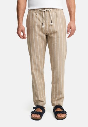 Mand iført beige, stribede hørbukser med snøre og sorte sandaler med korksåler, stående mod en ensfarvet baggrund.