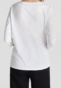 Witte langwerpige top met een wijde halslijn, lichte textuur en een ontspannen pasvorm, gecombineerd met zwarte onderkleding. Eenvoudig en veelzijdig ontwerp.