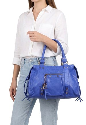 Frau trägt weißes Hemd und helle Jeans und hält eine mittelgroße blaue Lederhandtasche mit Fronttasche und dekorativen Quasten.