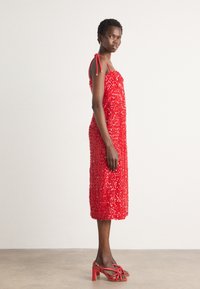 Robe sans manches rouge avec un design texturé, ornée de sequins. Elle dispose de bretelles ajustables et d'une coupe mi-genoux, accompagnée de sandales à talons rouges.